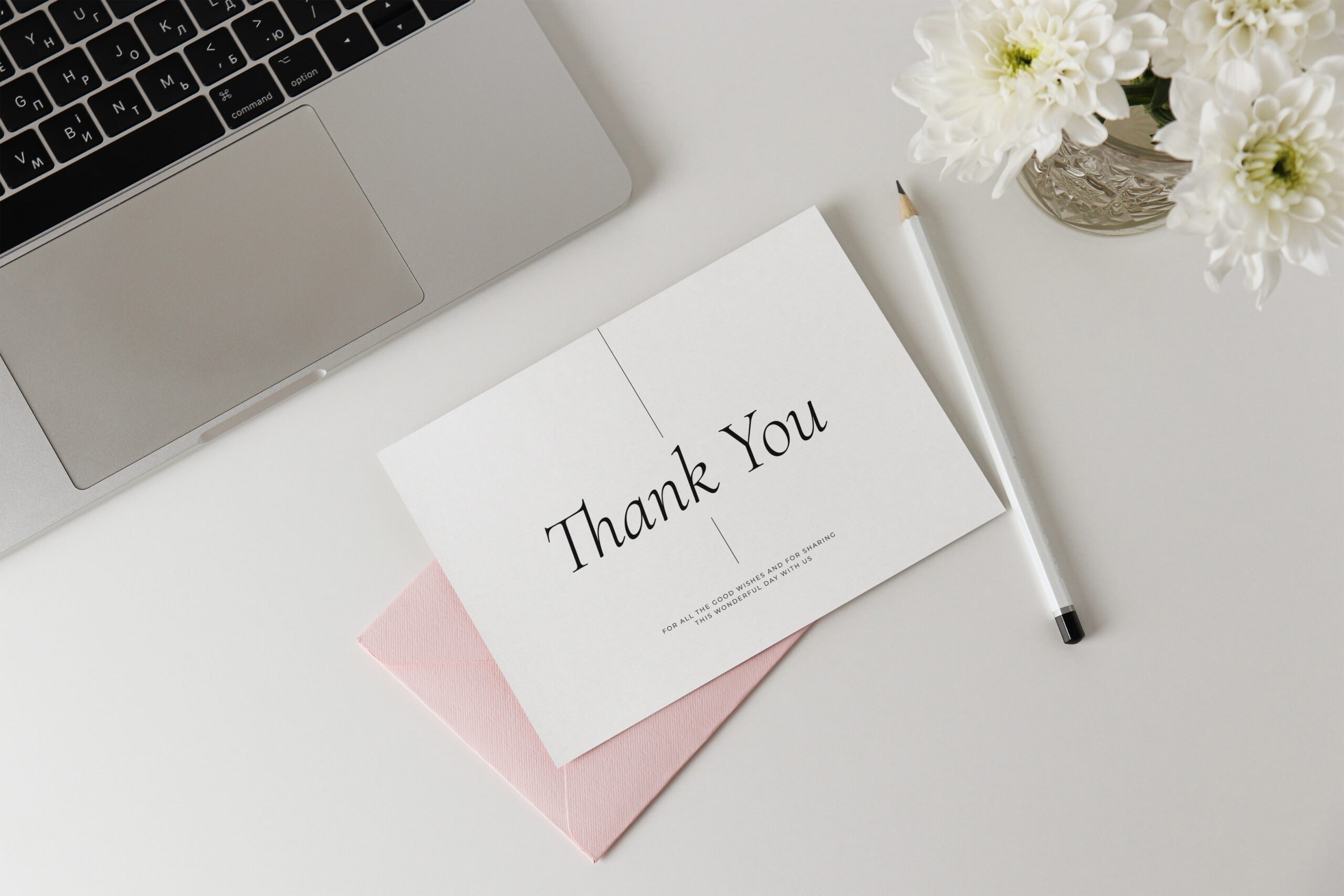 Photo of a thank you card with a pink envelope behind it on a white desk, with a laptop sitting open nearby and a pencil beside the card. There's a vase with white flowers nearby.