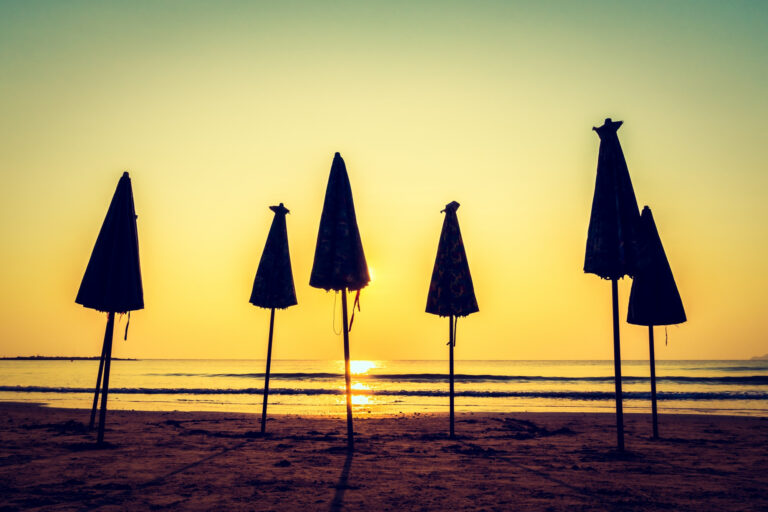 Photo of a beach at sunset with several closed umbrellas standing in the sand.
