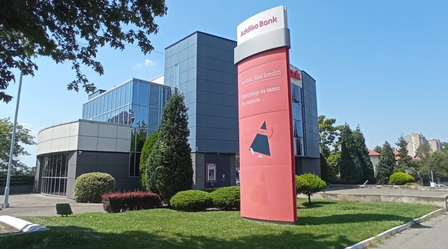 Photo of Addiko Bank - a large glass building with a tall red sign in front and lush green landscaping.