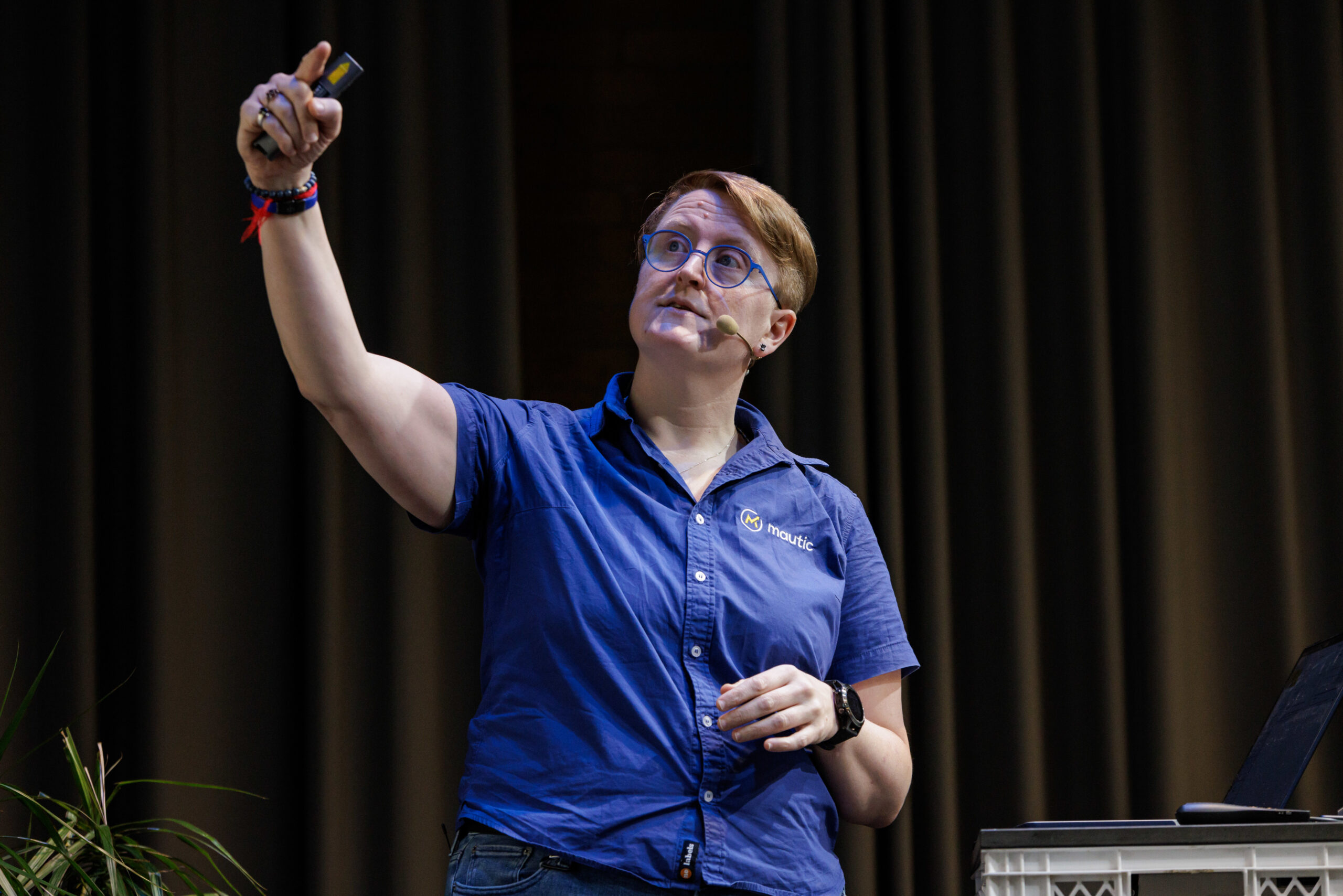 Photo of a white woman with short blonde hair on the stage at a conference wearing a blue collared shirt with short sleeves. She's holding a remote presenter in her left hand and pointing to a screen with the right hand.