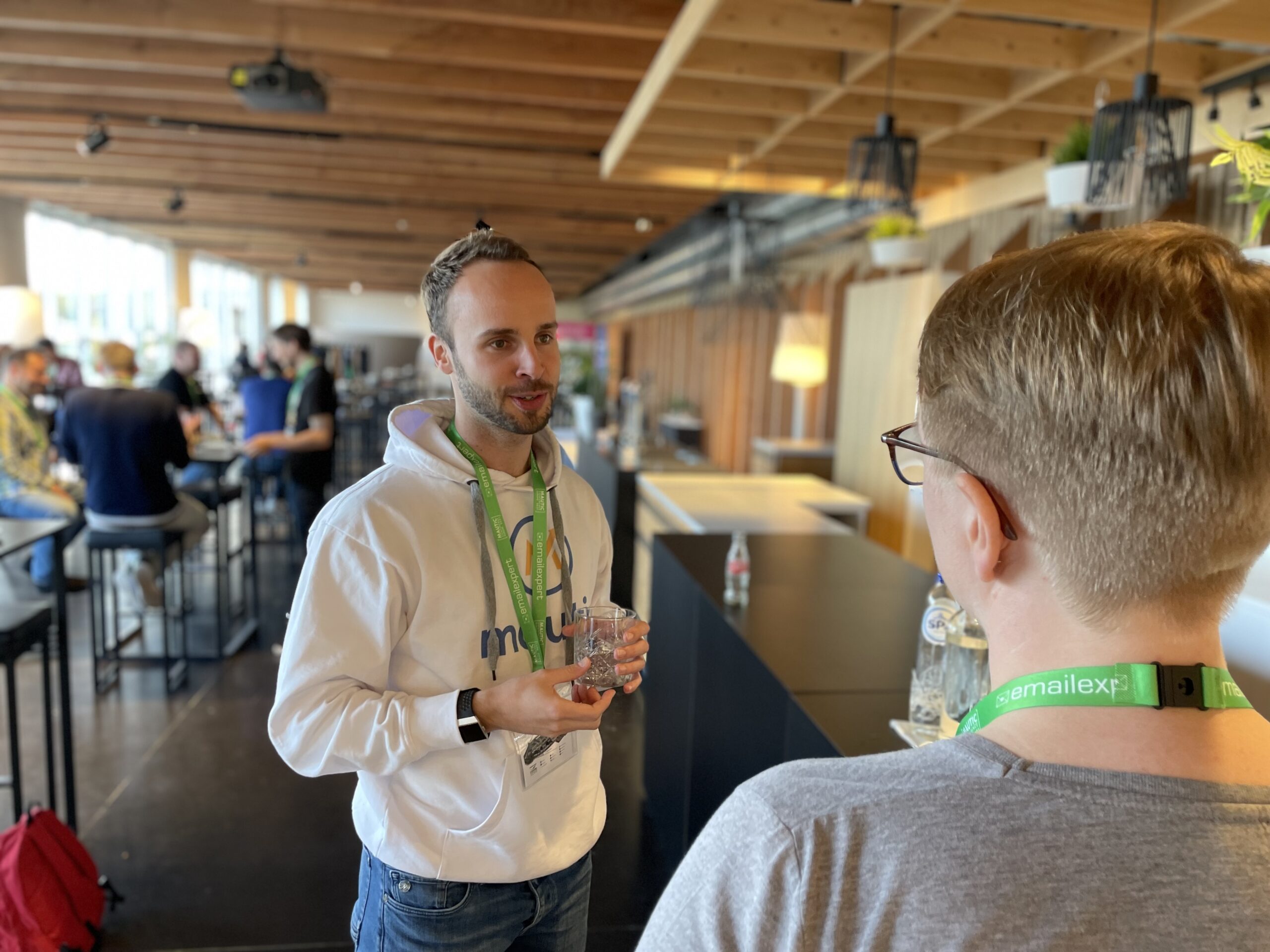 Photo of a woman with short hair from over her shoulder, talking to a young man standing holding a small glass tumbler wearing a white Mautic hoodie and blue jeans in a venue bar.