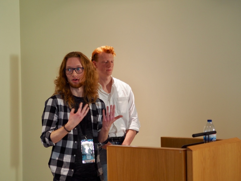 Photo of two men standing behind a lecturn, one has long ginger curly hair and the other has short ginger curly hair.