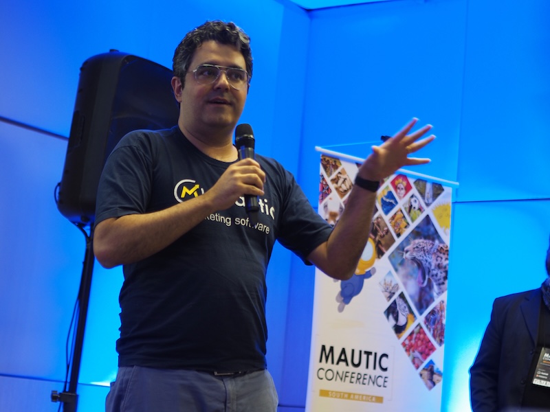Photo of a Brazilian man standing on a stage wearing a dark tshirt with Mautic marketing software on the front. He's holding a microphon and the background is illuminated in blue, with a speaker behind him and a popup with Mautic Conference to the right. He's wearing jeans and has glasses with short dark curly hair.