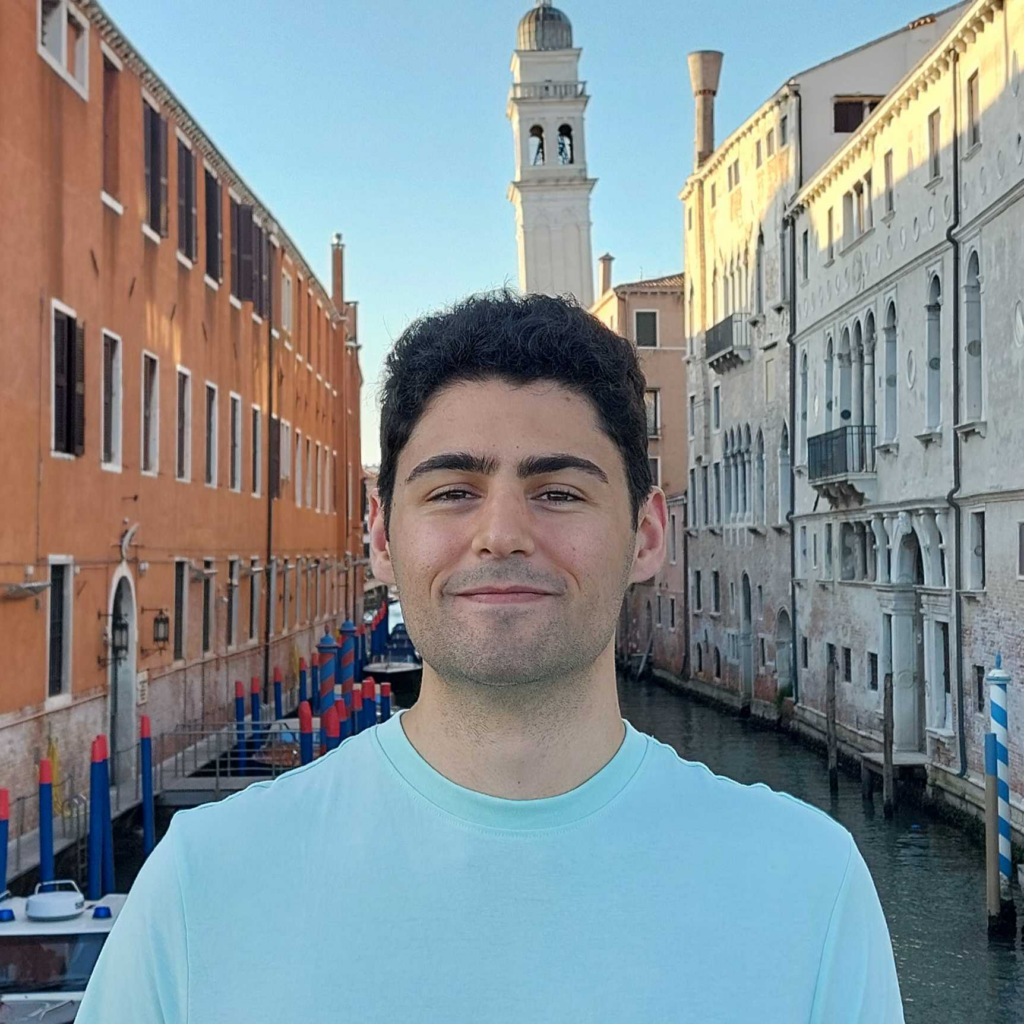 Photo of a Brazilian man with short, dark hair and a full beard and moustache standing in a park with tall skyscrapers behind him. He's wearing a red jacket and a blue coloured tshirt.